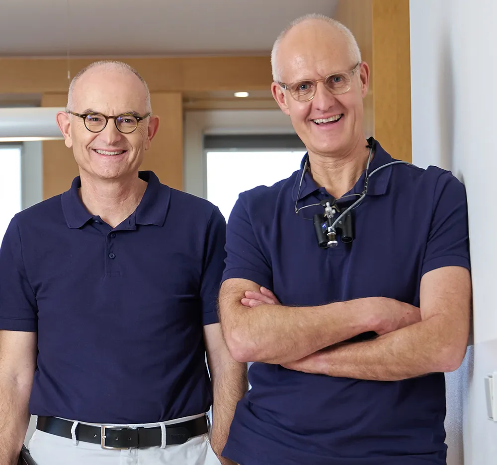 Das Zahnärzte-Team Dr. Frank MSc. MSc. und Dr. Pflumm stehen im Empfangsbereich der Zahnarztpraxis.