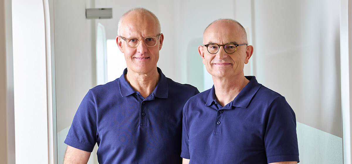 Die Zahnärzte Dr. Frank und Dr. Pflumm stehen nebeneinander in ihrer Praxis