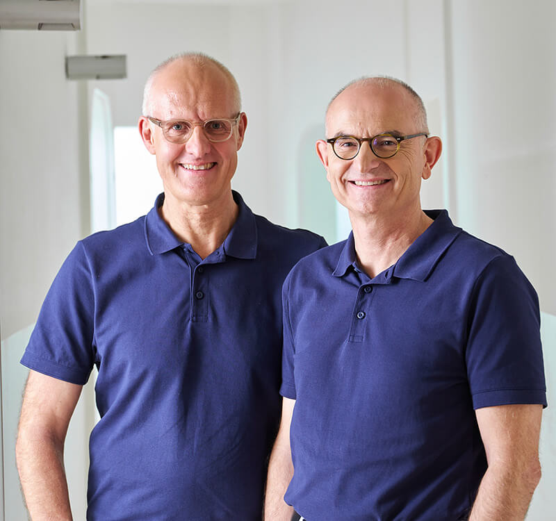 Die Zahnärzte Dr. Frank und Dr. Pflumm stehen nebeneinander in ihrer Praxis