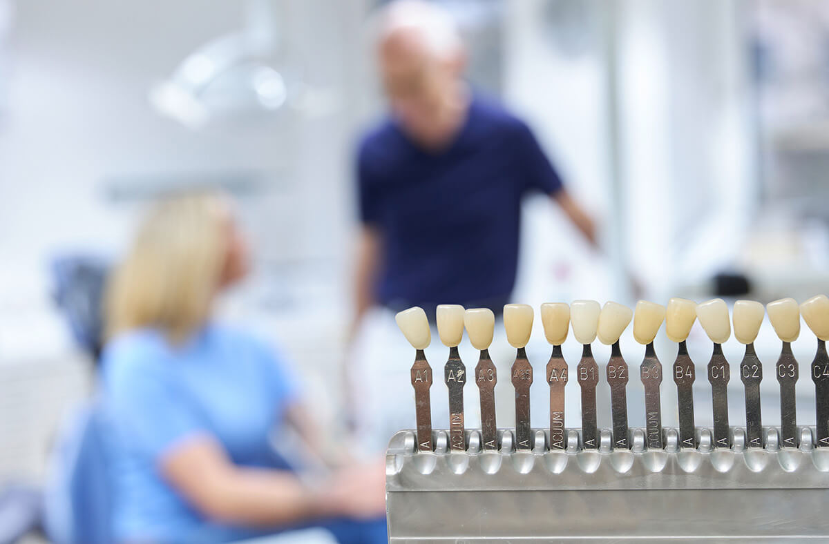 Hintergrund Dr. Frank und Patientin, Vordergrund Zahnweißkarte für Zahnfarbe bei Bleaching
