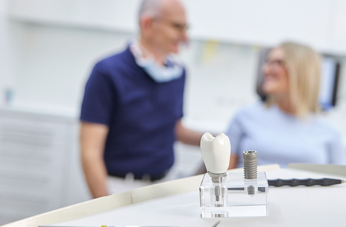 Dr. Frank mit einer Patientin im Hintergrund, ein Implantat-Modell im Vordergrund
